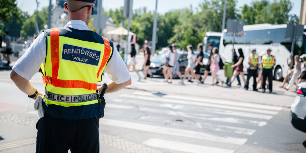 Egy forgalmas budapesti kereszteződésben magas láthatóságú, sárga‑narancssárga mellényt viselő rendőr áll a gyalogátkelőnél, háttal a kamerának. A zebrán több gyalogos halad át, a háttérben pedig két további egyenruhás rendőr áll egy fehér autóbusz mellett. A környezet városi: járművek, közlekedési táblák és zöld növényzet látható.