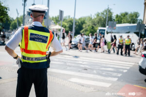 Egy forgalmas budapesti kereszteződésben magas láthatóságú, sárga‑narancssárga mellényt viselő rendőr áll a gyalogátkelőnél, háttal a kamerának. A zebrán több gyalogos halad át, a háttérben pedig két további egyenruhás rendőr áll egy fehér autóbusz mellett. A környezet városi: járművek, közlekedési táblák és zöld növényzet látható.