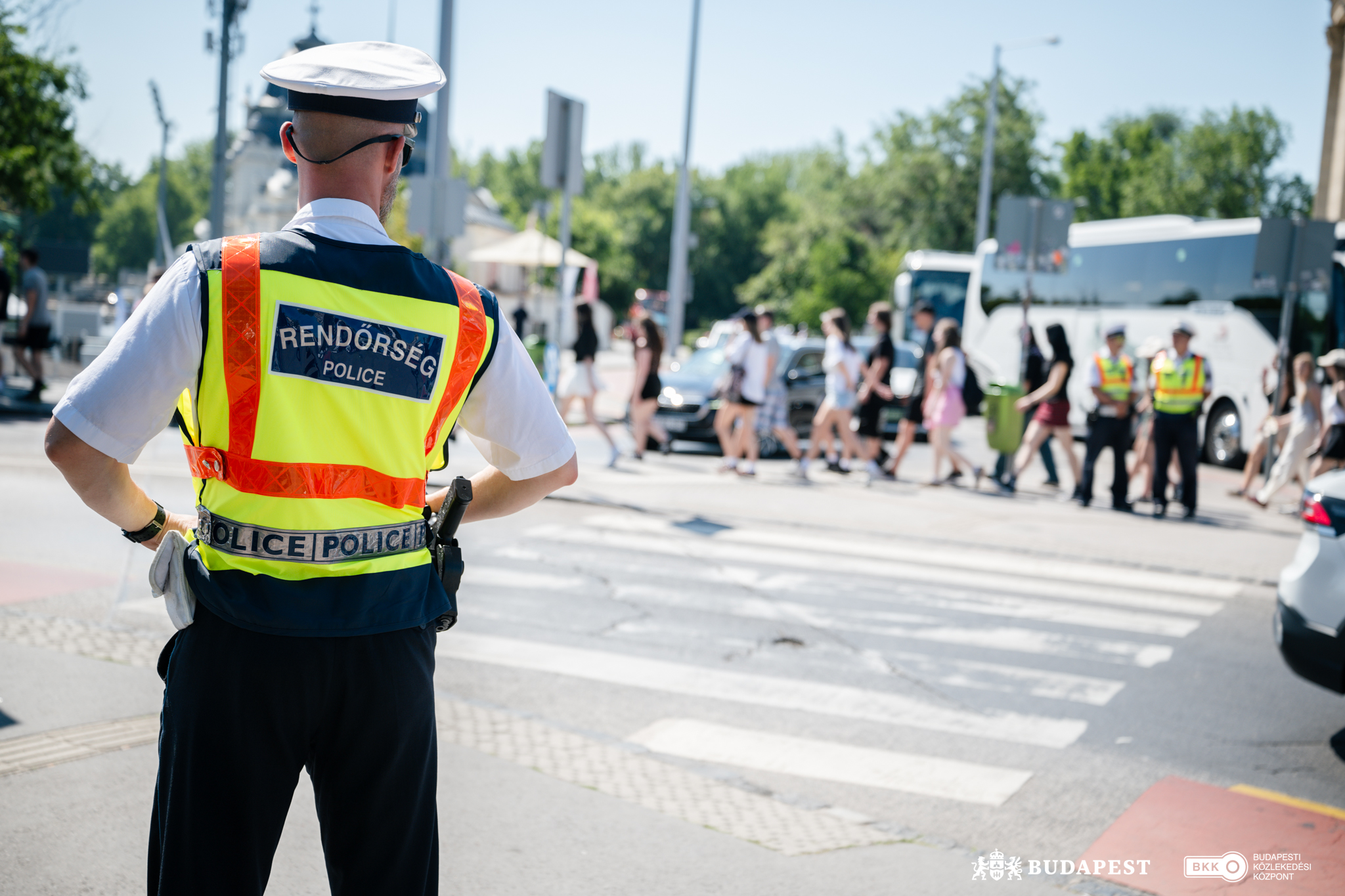 Egy forgalmas budapesti kereszteződésben magas láthatóságú, sárga‑narancssárga mellényt viselő rendőr áll a gyalogátkelőnél, háttal a kamerának. A zebrán több gyalogos halad át, a háttérben pedig két további egyenruhás rendőr áll egy fehér autóbusz mellett. A környezet városi: járművek, közlekedési táblák és zöld növényzet látható.