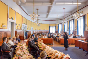 A polgármesteri hivatal dísztermében tartott ünnepségen a résztvevők sorokban ülve hallgatják Auth Csilla eMeRTon-díjas énekes előadását. A nagy, magas mennyezetű teremben fa burkolat, sárga falak és kék függönyök láthatók, a mennyezetről gömb alakú csillárok lógnak. Az asztalokon virágcsokrok sorakoznak, a közönség pedig figyelmesen a fellépő felé fordul.