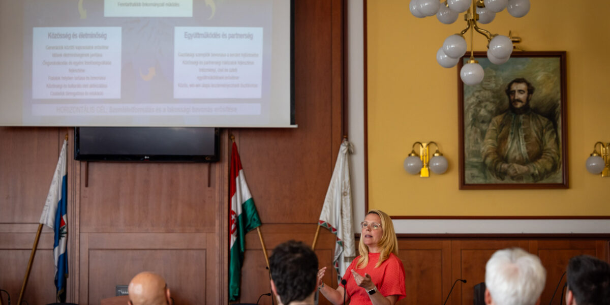 A polgármesteri hivatal tanácstermében tartott megbeszélésen a Civil Impact Nonprofit Kft. szakértői mutatják be a XV. kerület készülő fenntarthatósági stratégiáját a képviselő‑testület tagjainak. A terem elején álló előadó a kivetítőn megjelenő prezentációra hivatkozik, miközben a résztvevők sorokban ülve figyelik a tájékoztatást. A háttérben a magyar zászló és egy nagy méretű portré látható, a falakon fa burkolat fut végig.