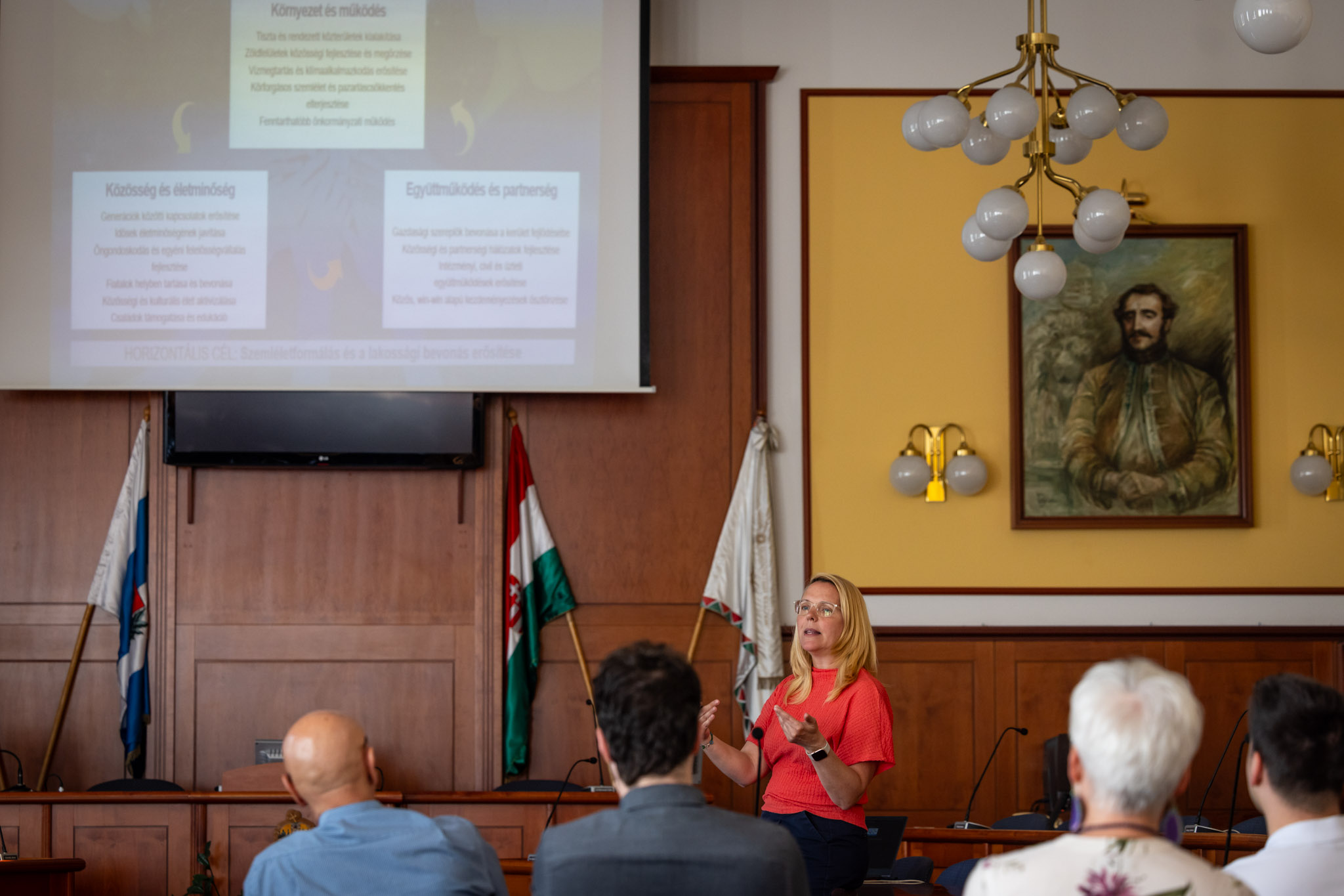 A polgármesteri hivatal tanácstermében tartott megbeszélésen a Civil Impact Nonprofit Kft. szakértői mutatják be a XV. kerület készülő fenntarthatósági stratégiáját a képviselő‑testület tagjainak. A terem elején álló előadó a kivetítőn megjelenő prezentációra hivatkozik, miközben a résztvevők sorokban ülve figyelik a tájékoztatást. A háttérben a magyar zászló és egy nagy méretű portré látható, a falakon fa burkolat fut végig.