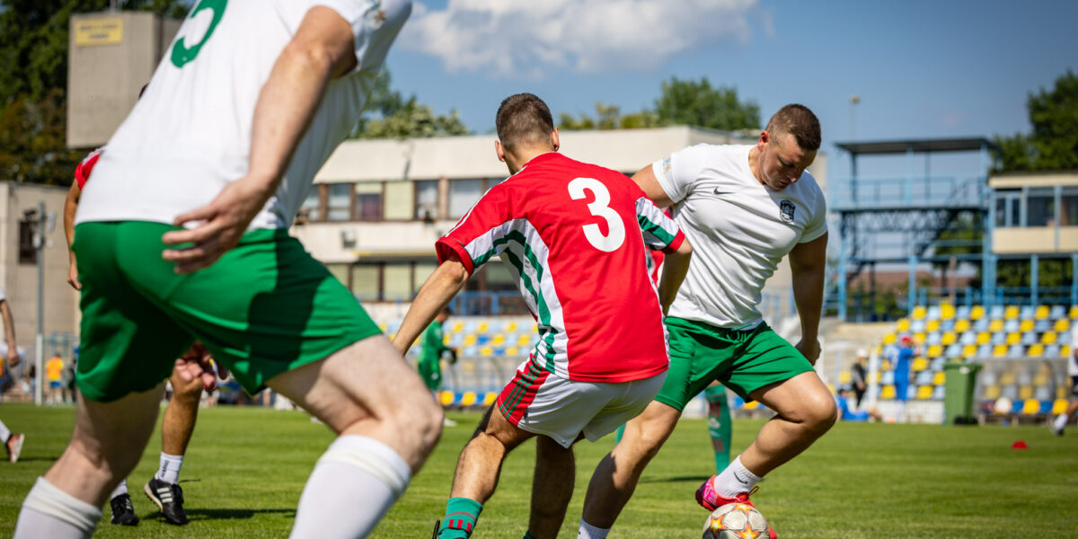 Egy focimeccs jelenete a Budai II László Stadion gyepén. A kép előterében egy piros mezben játszó játékos próbálja szerelni a labdát vezető, fehér mezű ellenfelét, miközben egy másik fehér mezű játékos közelről követi az akciót. A háttérben a stadion lelátója és az épületek láthatók, napos időben.