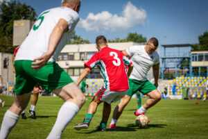 Egy focimeccs jelenete a Budai II László Stadion gyepén. A kép előterében egy piros mezben játszó játékos próbálja szerelni a labdát vezető, fehér mezű ellenfelét, miközben egy másik fehér mezű játékos közelről követi az akciót. A háttérben a stadion lelátója és az épületek láthatók, napos időben.