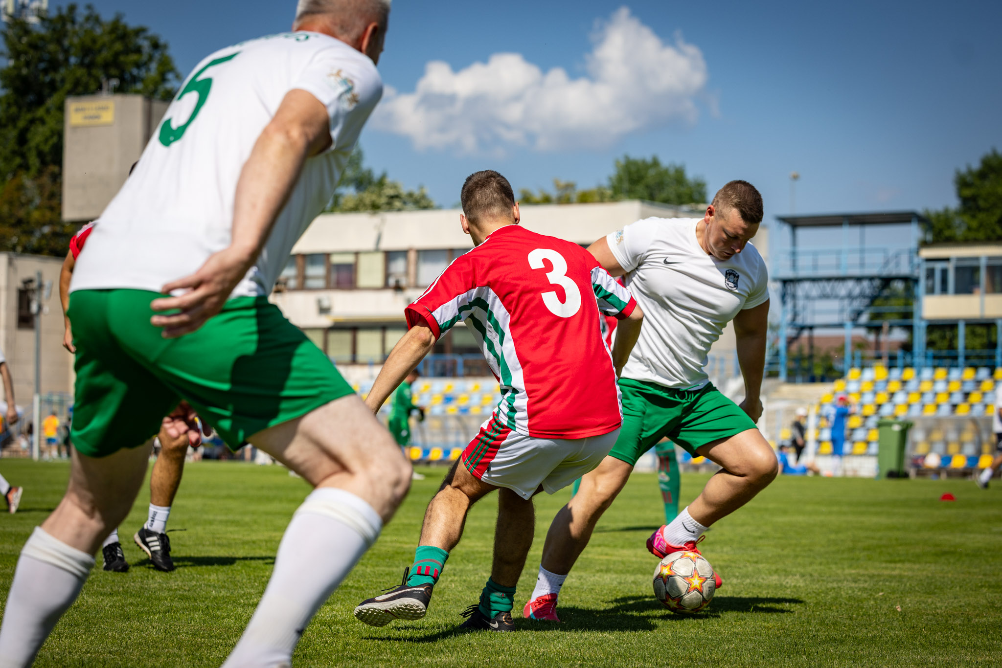 Egy focimeccs jelenete a Budai II László Stadion gyepén. A kép előterében egy piros mezben játszó játékos próbálja szerelni a labdát vezető, fehér mezű ellenfelét, miközben egy másik fehér mezű játékos közelről követi az akciót. A háttérben a stadion lelátója és az épületek láthatók, napos időben.