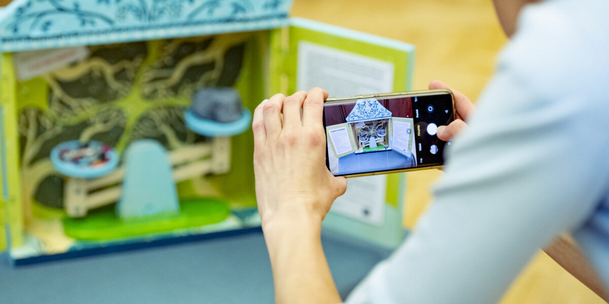 Egy fiatal a telefonjával fotóz egy asztalon álló, színes, miniatűr diorámát. A kis makett ház- vagy színpadszerű szerkezet, kék és zöld mintákkal díszítve. A belső térben apró tárgyak – például egy pad és egy kalap – láthatók. A jelenet a kreatív alkotást és annak dokumentálását mutatja be.
