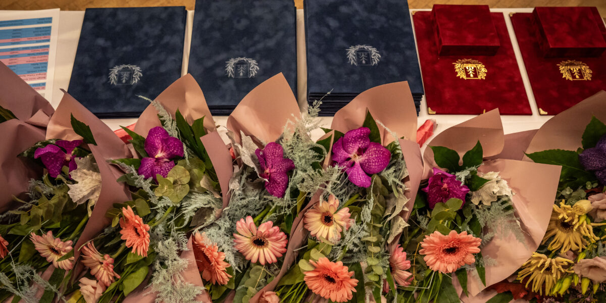 Egy terített asztalon több csokor friss virág és egymásra rendezett, díszes oklevélmappák láthatók. A virágcsokrok rózsaszín gerberákat, lila orchideákat és zöld leveleket tartalmaznak, halvány rózsaszín papírba csomagolva. Mögöttük két nagyobb halomban kék, mellettük kisebb számban bordó oklevélmappák sorakoznak, mindegyiken arany színű címerrel. A kompozíció ünnepi hangulatot áraszt, és egy közelgő kerületi elismerés vagy díszpolgári díjátadó előkészületeit idézi.