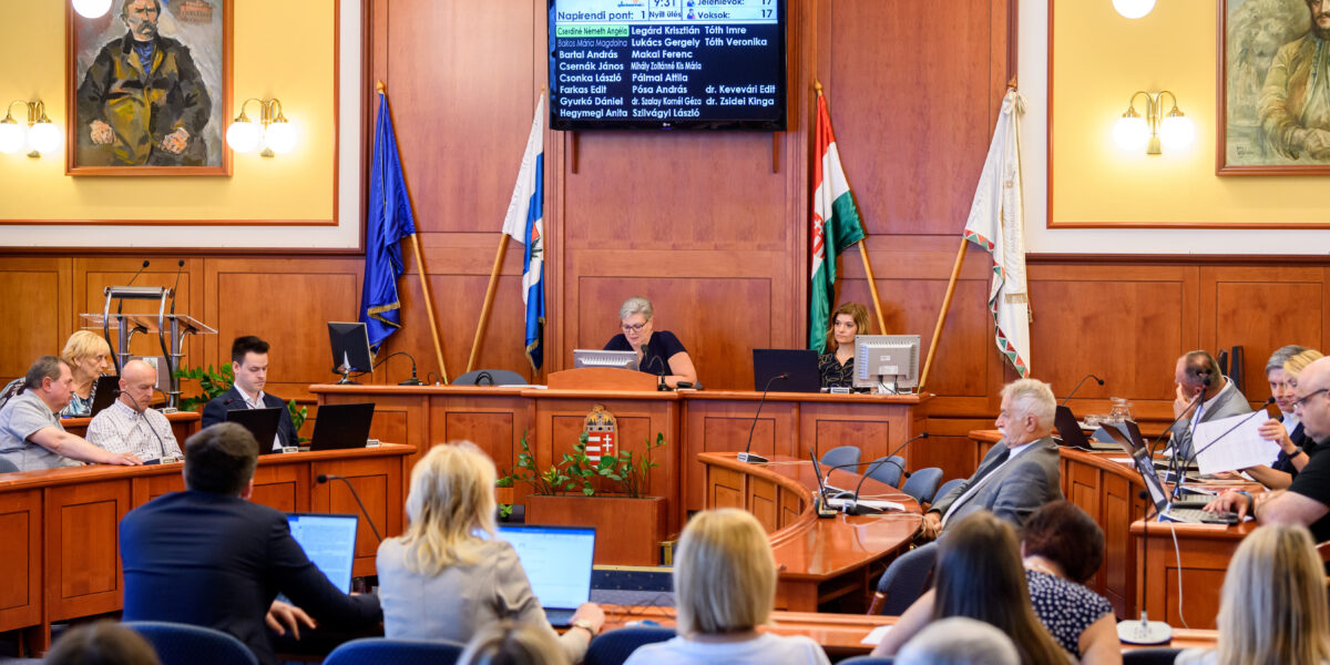 A XV. kerületi Képviselő-testület ülésterme látható a képen, fa burkolatú falakkal, csillárokkal és a falon elhelyezett portrékkal. A terem közepén nagy, íves tárgyalóasztal körül ülnek a képviselők és a hivatal munkatársai, előttük laptopok, iratok és jegyzetek. A terem elején, emelt pódiumon a polgármester és a jegyző dolgozik számítógépnél, mögöttük a magyar zászló és további önkormányzati zászlók állnak. Fölöttük digitális kijelzőn a napirendi pont és a jelenlévő képviselők nevei olvashatók. A résztvevők figyelmesen követik az ülést, többen jegyzetelnek vagy a monitorukat nézik, a jelenet egy hivatalos, döntéshozatali folyamat pillanatát rögzíti.