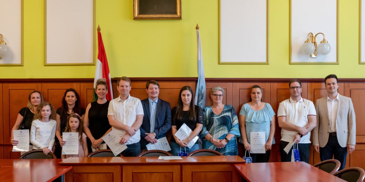 A Polgármesteri Hivatal termében tizenegy ember áll egymás mellett egy hosszú, fából készült tárgyalóasztal mögött. Mindannyian oklevelet tartanak a kezükben. A csoport közepén Cserdiné Németh Angéla polgármester látható, kék blúzban. Tőle jobbra, a sor végén áll Csernák János alpolgármester. A háttérben sárga falak, fa falburkolat, két zászló (a magyar nemzeti zászló és egy világoskék önkormányzati zászló), valamint egy falra akasztott portré látható. A jelenet a közösségi költségvetés nyertes ötletbeadóinak ünnepélyes köszöntését örökíti meg.