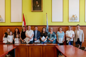 A Polgármesteri Hivatal termében tizenegy ember áll egymás mellett egy hosszú, fából készült tárgyalóasztal mögött. Mindannyian oklevelet tartanak a kezükben. A csoport közepén Cserdiné Németh Angéla polgármester látható, kék blúzban. Tőle jobbra, a sor végén áll Csernák János alpolgármester. A háttérben sárga falak, fa falburkolat, két zászló (a magyar nemzeti zászló és egy világoskék önkormányzati zászló), valamint egy falra akasztott portré látható. A jelenet a közösségi költségvetés nyertes ötletbeadóinak ünnepélyes köszöntését örökíti meg.