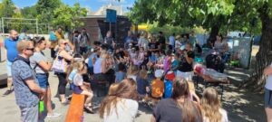 Ozmán utcai Szomszédünnep kisállatbemutatója. A park árnyas fái alatt gyerekek és felnőttek ülnek körben, előttük egy előadó mutat be kisállatokat egy asztalnál elhelyezett dobozokból. A háttérben játszótér és parképületek láthatók, barátságos, közösségi hangulatban.