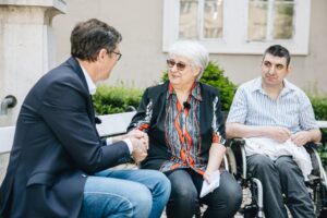 Egy kültéri padon három ember beszélget. Karácsony Gergely főpolgármester a bal oldalon ül, és kezet fog a mellette ülő Schilling Magdolnával, a Gondoskodás Gyermekeinkért Alapítvány elnökével. Jobbra tőlük Fecó ül egy kerekesszékben, figyelve a beszélgetést. A háttérben ablakos épület és udvari növényzet látható.