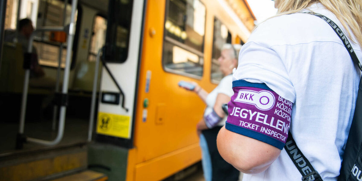 A fotón egy budapesti villamosmegállóban zajló jegyellenőrzés látható. A kép előterében egy fehér pólót viselő jegyellenőr áll, bal karján jól látható a lila színű karszalag, rajta a felirat: „BKK JEGYELLENŐR – TICKET INSPECTOR”. A jegyellenőr a villamos nyitott ajtaja mellett helyezkedik el, testtartása arra utal, hogy éppen az utasok felszállását vagy jegyeinek ellenőrzését figyeli. A háttérben egy másik BKK‑jegyellenőr is látható, kezében ellenőrző készülékkel. A villamos ajtaja nyitva áll, a sárga fellépő és a fehér ajtókeret jól kivehető. A jármű belsejében egy utas körvonala is látszik, amint a kocsiban tartózkodik. A jelenet egy forgalmas, hétköznapi pillanatot rögzít a budapesti közösségi közlekedésben, ahol a jegyellenőrök a szabályos utazást és az utasbiztonságot felügyelik.