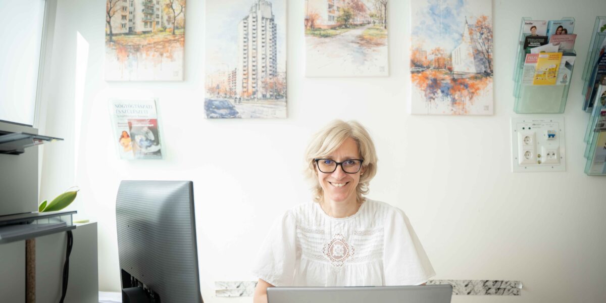 Dr. Fekete Márta egy világos, rendezett rendelőben ül az íróasztalnál. Előtte laptop és monitor, körülötte tollak, papírok és irodai eszközök. A falon több, Újpalotát ábrázoló kerületi fotó látható keretben, mellettük fali tartókban egészségügyi kiadványok. A doktornő koncentráltan dolgozik, a helyiségbe balról természetes fény áramlik be.