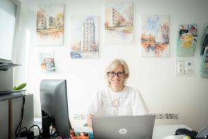 Dr. Fekete Márta egy világos, rendezett rendelőben ül az íróasztalnál. Előtte laptop és monitor, körülötte tollak, papírok és irodai eszközök. A falon több, Újpalotát ábrázoló kerületi fotó látható keretben, mellettük fali tartókban egészségügyi kiadványok. A doktornő koncentráltan dolgozik, a helyiségbe balról természetes fény áramlik be.