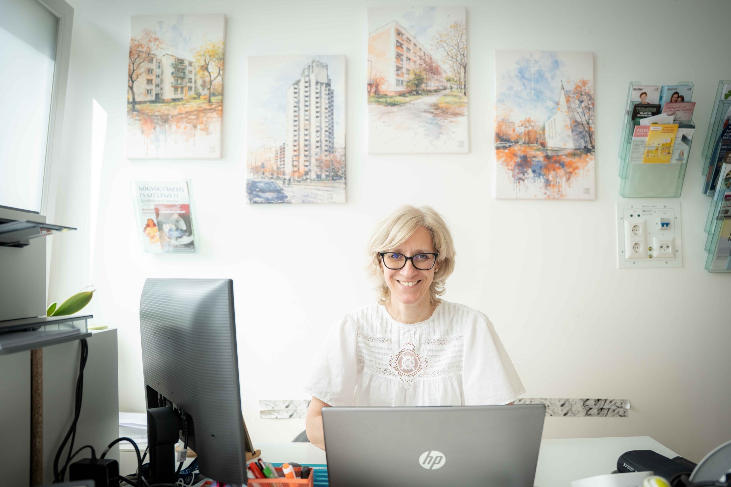 Dr. Fekete Márta egy világos, rendezett rendelőben ül az íróasztalnál. Előtte laptop és monitor, körülötte tollak, papírok és irodai eszközök. A falon több, Újpalotát ábrázoló kerületi fotó látható keretben, mellettük fali tartókban egészségügyi kiadványok. A doktornő koncentráltan dolgozik, a helyiségbe balról természetes fény áramlik be.
