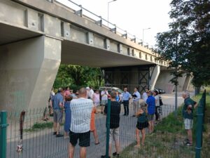 Drégelyvár utcai felüljáró fórum. A felüljáró masszív betonpillérei alatt egy csoport lakos és önkormányzati vezető állva beszélget, mögöttük parkoló autók és kerítéssel határolt terület látható. A résztvevők a környezet rendezéséről egyeztetnek, miközben a háttérben fák és városi zöldfelület keretezi a helyszínt.